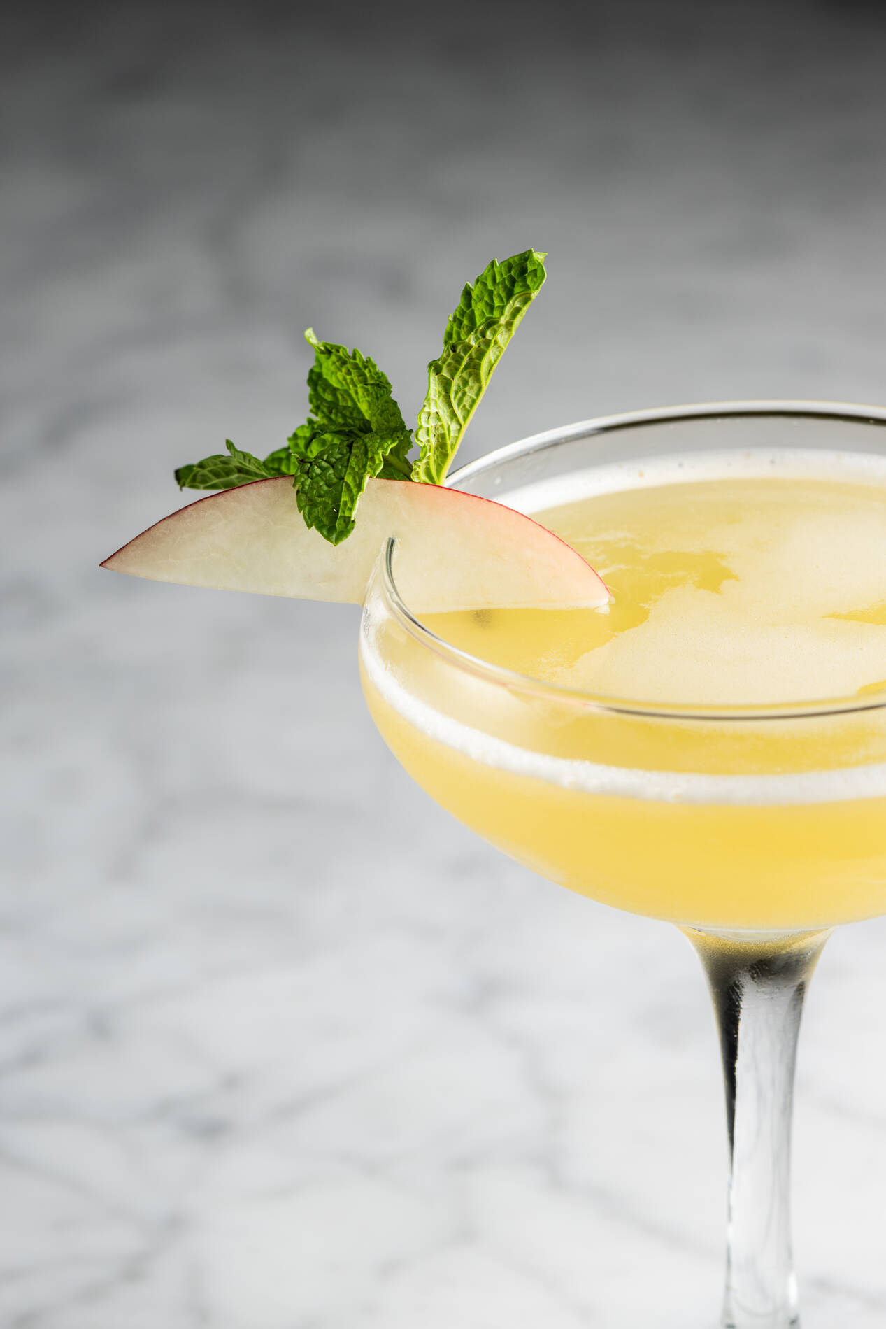 Close-up of a yellow cocktail in a coupe glass garnished with a mint sprig and apple slice.