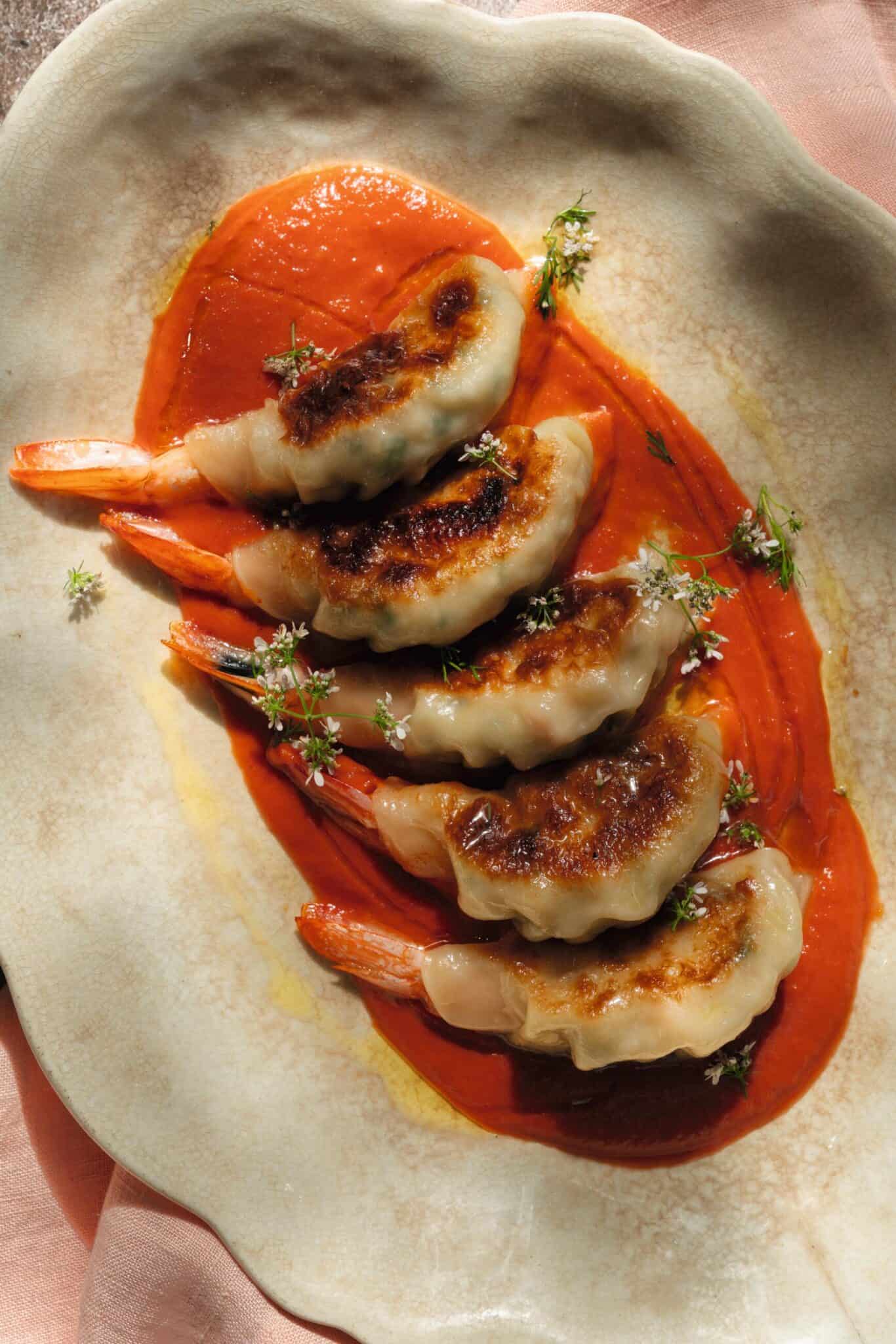 Five pan-seared shrimp dumplings served on a bed of red sauce, garnished with small white flowers.