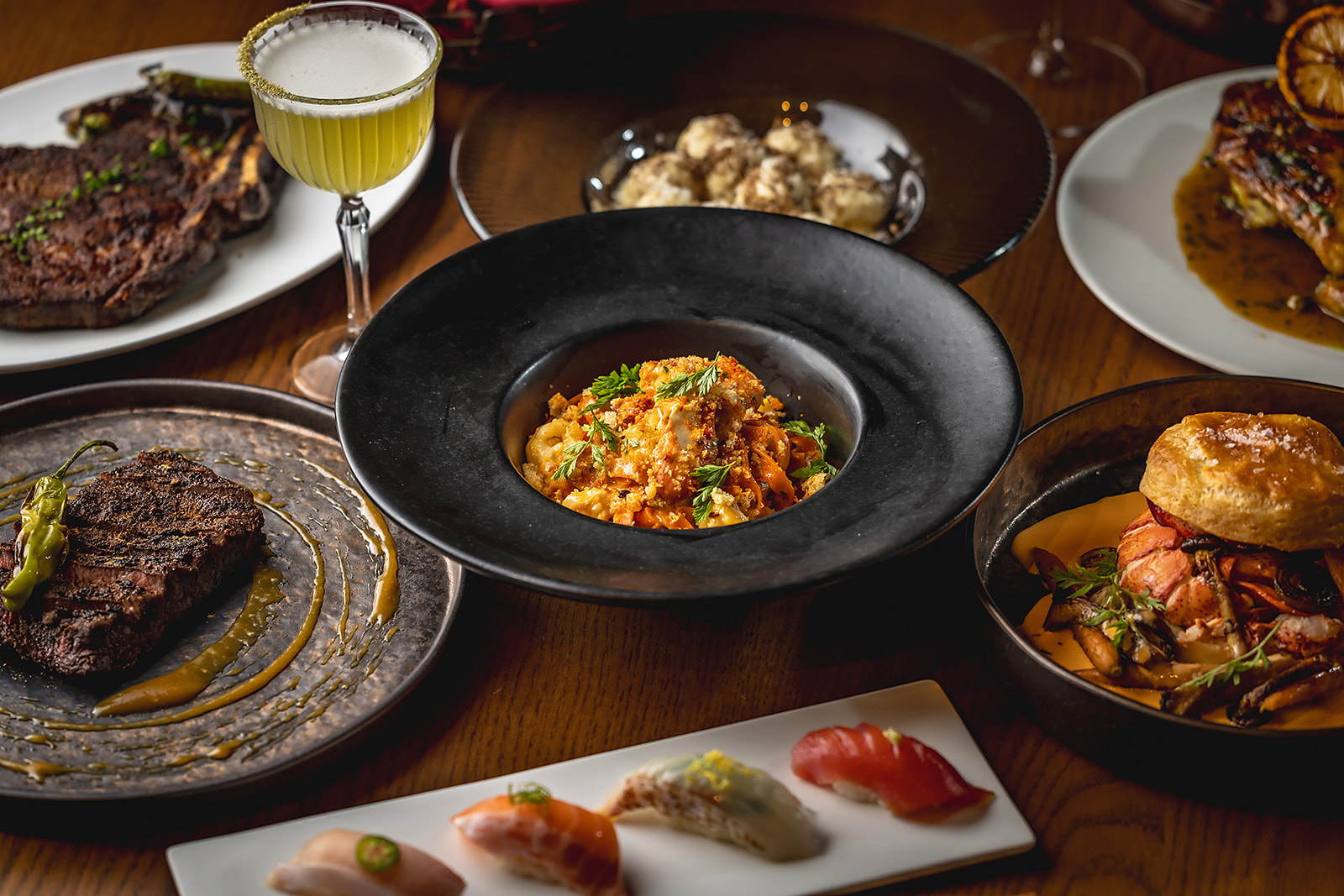 A spread of Carlyle Club dishes includes grilled steaks, lobster pot pie, sushi, pasta, sides, and a green cocktail.