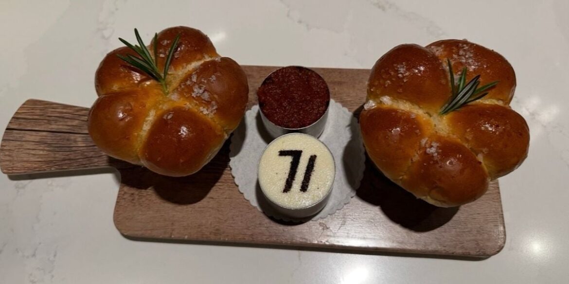 A delicious preview of Steakhouse 71 at Disney’s Contemporary Resort: two round rosemary-topped bread rolls and two mini desserts—one featuring the “71” logo—beautifully presented on a wooden board. Reservations are a must for this popular Walt Disney World dining spot.