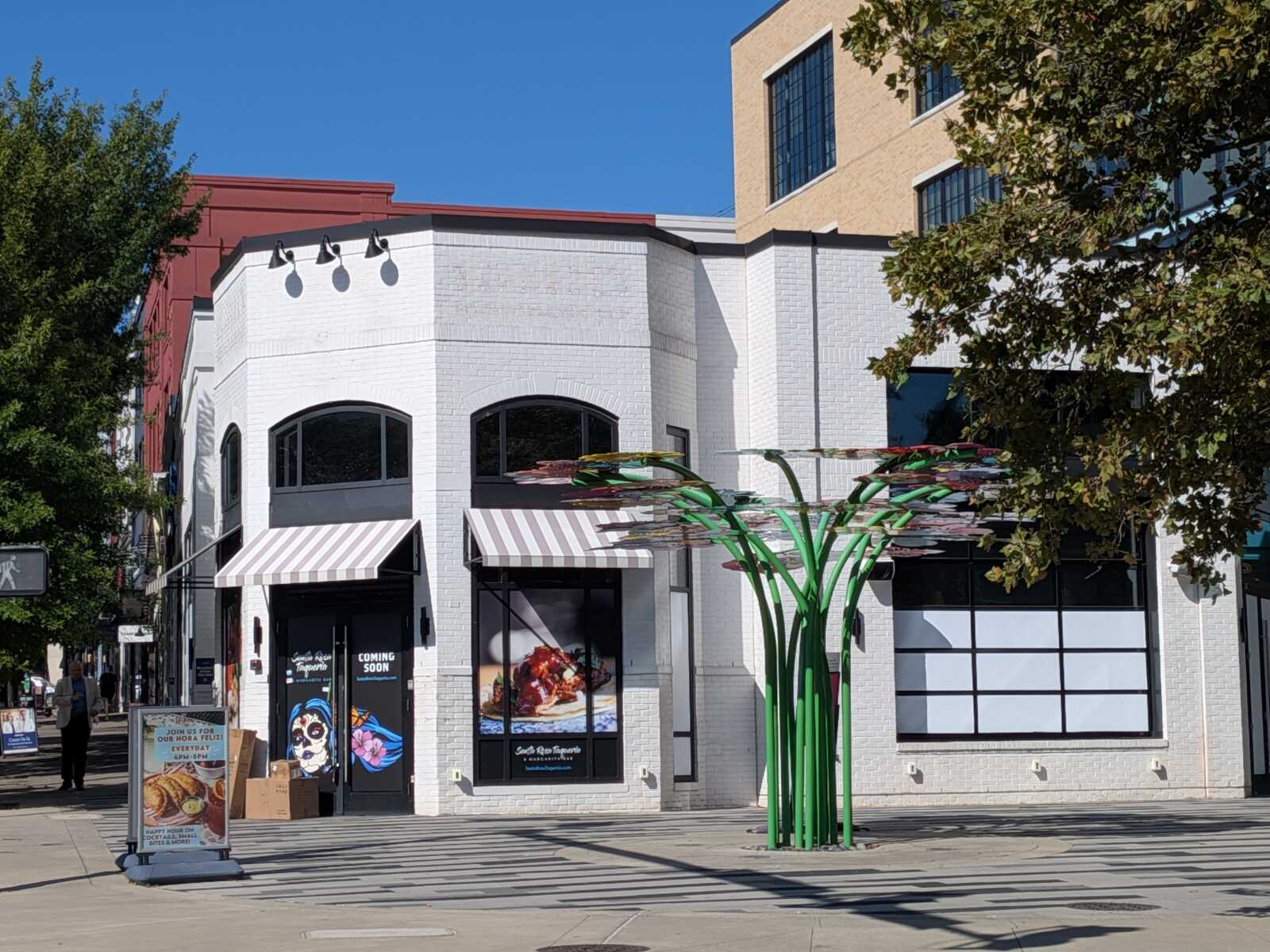 White brick storefronts with tan and white striped awnings on a sunny street; a green, sculptural tree-like installation in front.