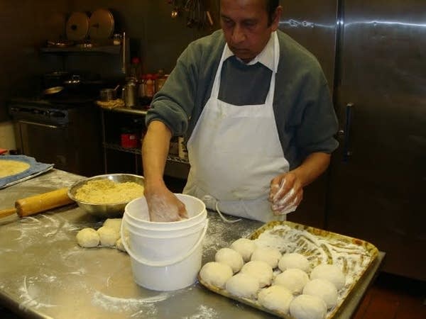 A man in a white apron reaches into a bucket while kneading dough on a kitchen counter.