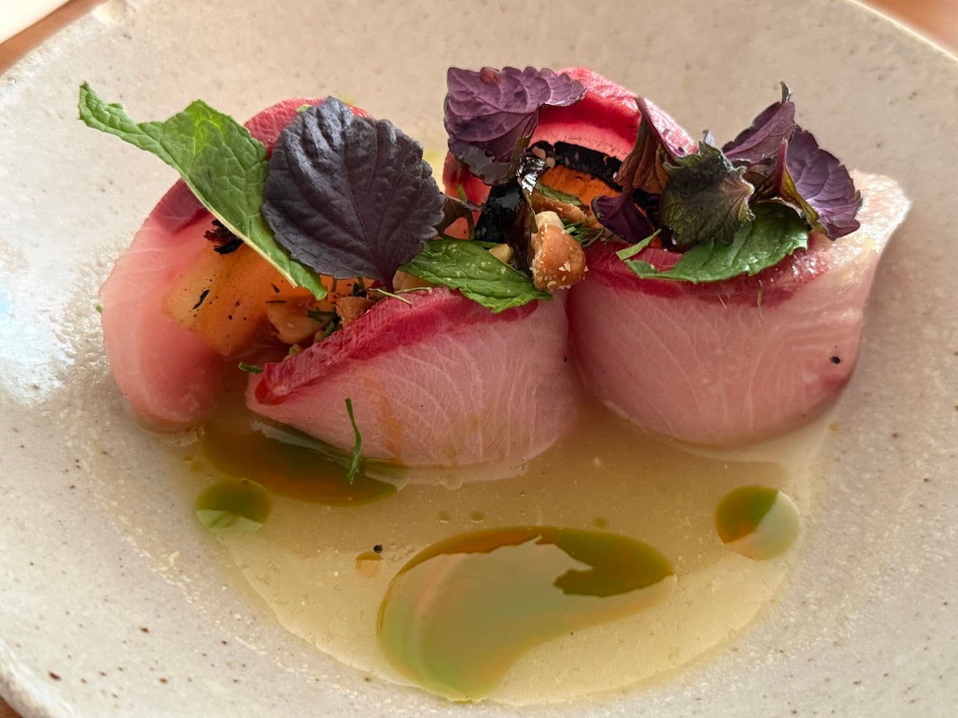 Two pieces of sashimi-style fish topped with fresh herbs and microgreens, served in a shallow bowl with light broth and green oil drizzles.