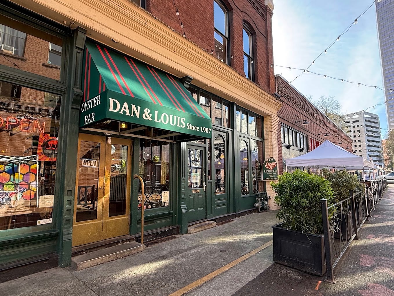 Street view of Dan & Louis Oyster Bar with green awning, outdoor seating under a white tent, string lights, and a historic brick building in an urban setting on a sunny day.