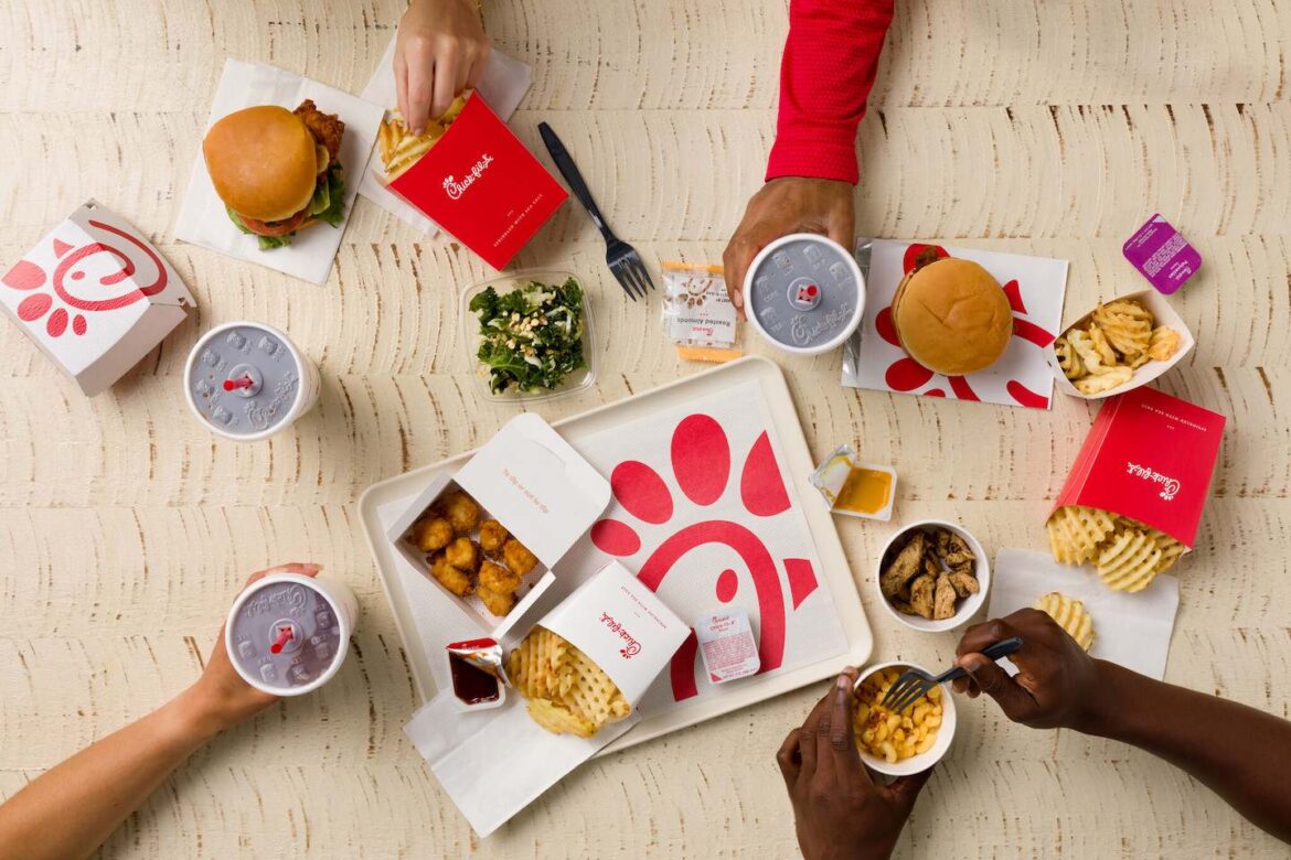 Chick-fil-A Announces New Cornelius Restaurant, Opening Oct. 8 Overhead view of Chick-fil-A items, including chicken sandwiches, fries, chicken nuggets, and drinks, on a wooden table with visible hands interacting with the food.