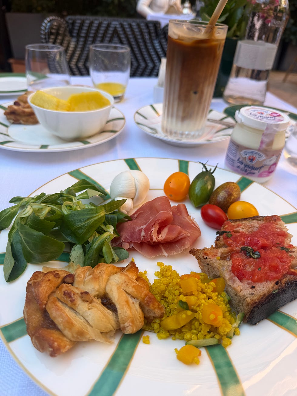 breakfast plate with salad, prosciutto, tomato bread, pastry, quinoa, and iced coffee.