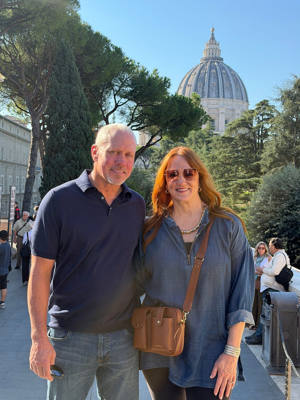 couple smiling in front of st. peter's basilica, surrounded by trees.