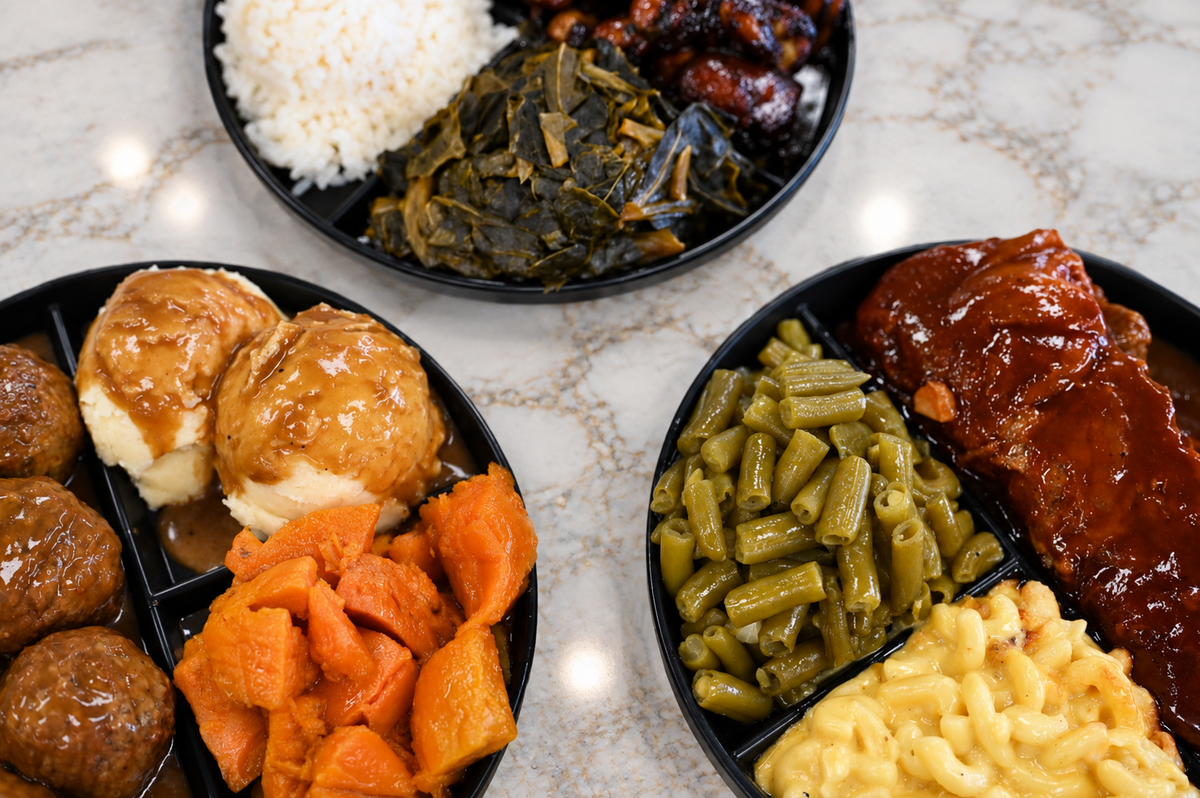 Top-down view of three black compartmentalized dinner plates containing soul food options: one with meatballs, mashed potatoes with gravy, and candied yams; another with BBQ ribs, green beans, and mac and cheese; and a third with dark meat BBQ chicken, collard greens, and white rice.