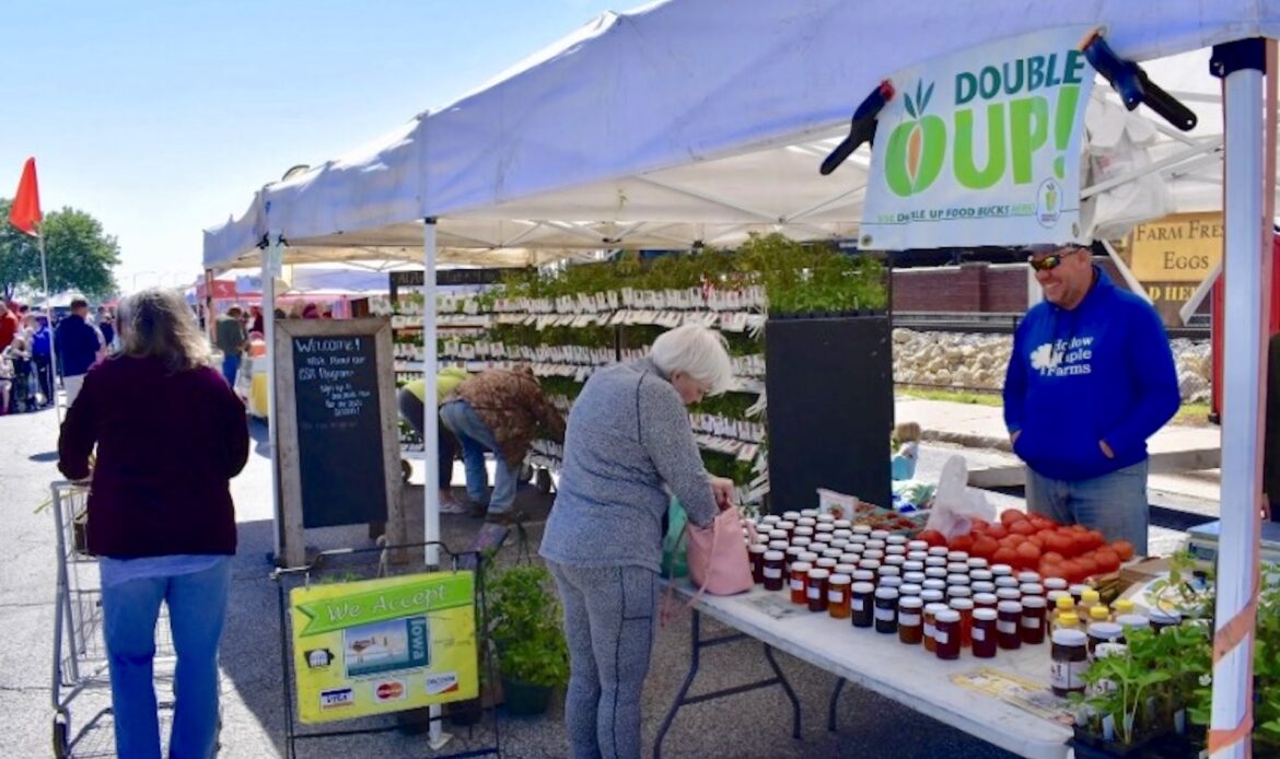 Michigan-born SNAP program turns $20 into $40 of produce at farmers markets A busy farmers' market with vendors displaying jars of preserves, vegetables, and plants under tents.