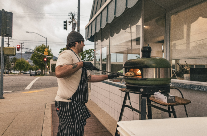 A person in an apron slides bread into a green outdoor pizza oven on a sidewalk.