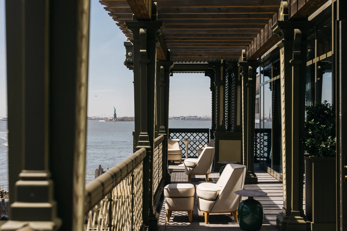 Rooftop terrace with seating, overlooking water and the Statue of Liberty in the distance under a clear sky.