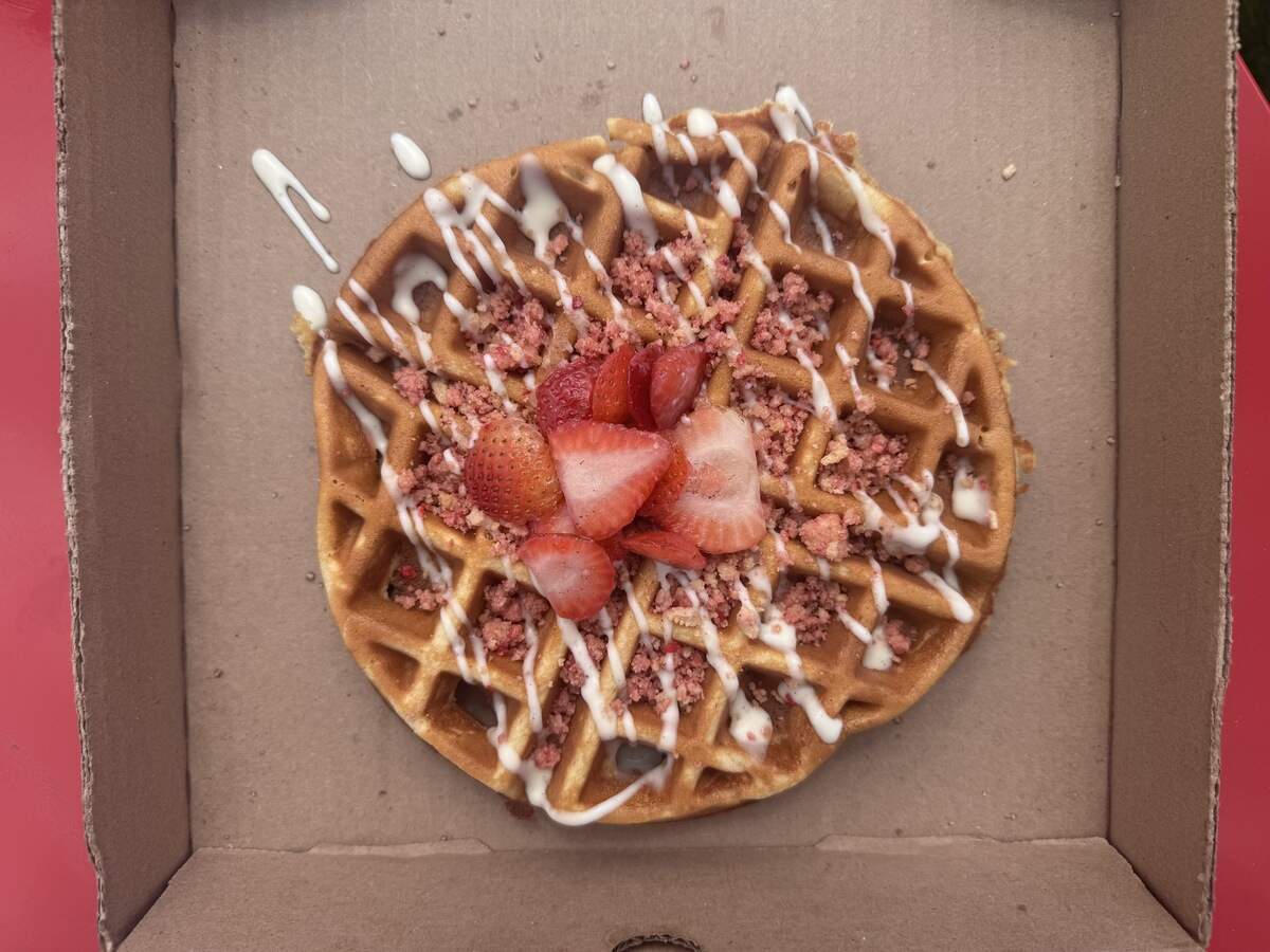 A golden-brown waffle inside a cardboard box, topped with sliced fresh strawberries, pink crumbles, and a drizzle of white icing, set against a red background.