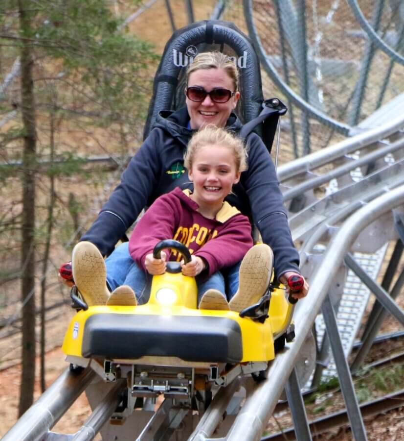 An adult and a child ride a yellow roller coaster together, both smiling, on a track through a wooded area—just one exciting way to play in Georgia.