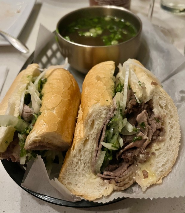 A sandwich with thinly sliced beef, greens and onions. There's a crock of dipping sauce with scallions and herbs on top in the background