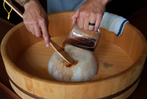 Pouring soy sauce over rice in a wooden barrel with a wooden paddle in the opposite hand.