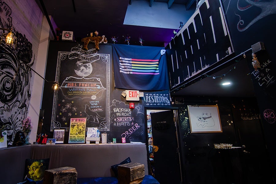 A cozy indoor space with dark walls decorated with various artworks and chalkboard signs. A large chalkboard features a moon illustration and text about