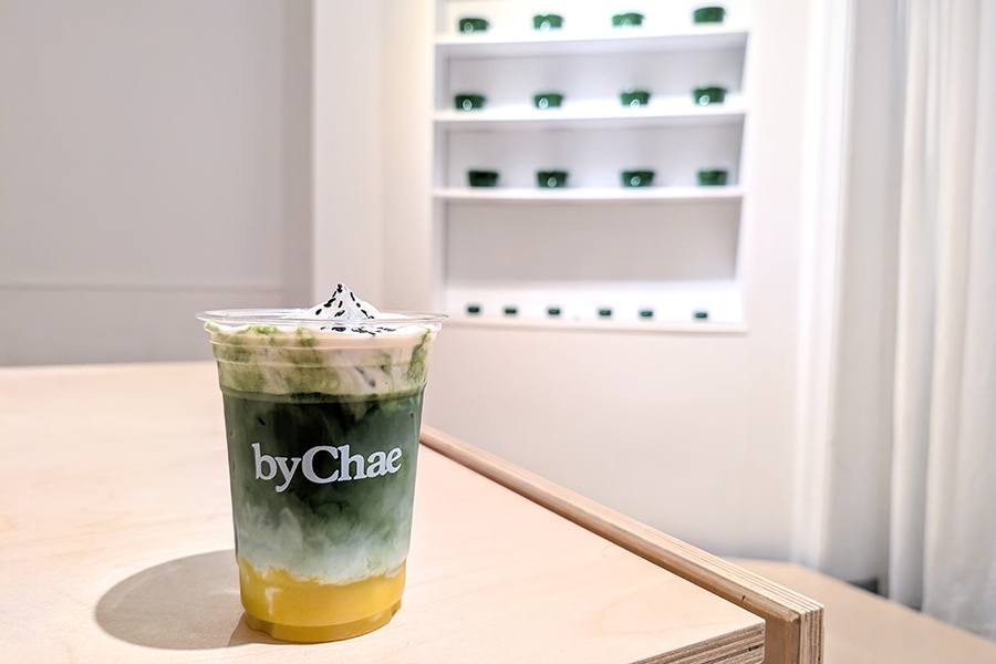 A plastic cup with a logo for the brand byChae is full of a layered matcha drink, with orange puree at the bottom, green matcha, and thick white foam. In the background, shelves hold green ceramic bowls.