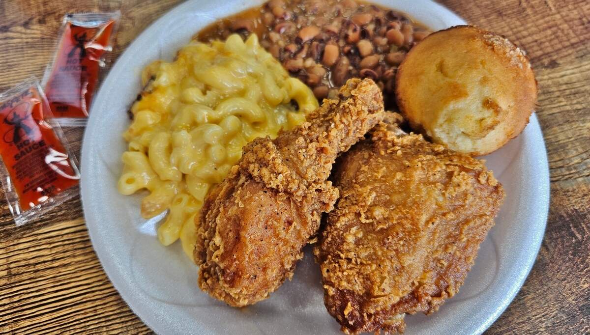 A close-up, overhead shot of a generous serving of Southern comfort food on a white disposable plate. Two pieces of crispy, golden-brown fried chicken (a drumstick and a thigh) are prominent in the foreground. Behind them, a large portion of creamy, cheesy macaroni and cheese is visible on the left, and a serving of brown-eyed peas or similar beans in a sauce is on the right. A cornbread muffin sits at the top right of the plate. Two packets of hot sauce are visible in the upper left corner of the frame. The plate rests on a wooden surface.