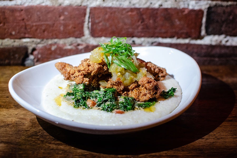 A white, shallow bowl contains a serving of creamy grits topped with crispy fried fish pieces, sautéed greens, and a dollop of chunky green relish or salsa, garnished with fresh green herbs. The dish is set on a wooden table with a brick wall background.