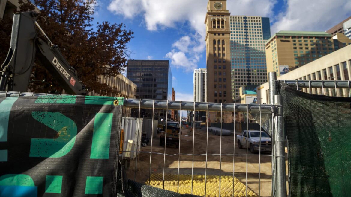 Denver restaurant says road work drove sales down 30% despite putting up its own detour signs A construction site in Denver with a fence, heavy machinery, and tall buildings in the background under a partly cloudy sky.