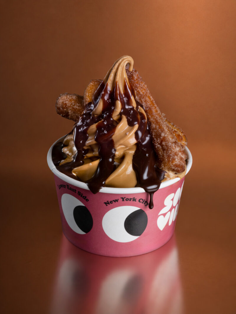 A swirl of Softville NYC soft-serve ice cream topped with chocolate syrup and two churros in a pink cup with a googly eye design.