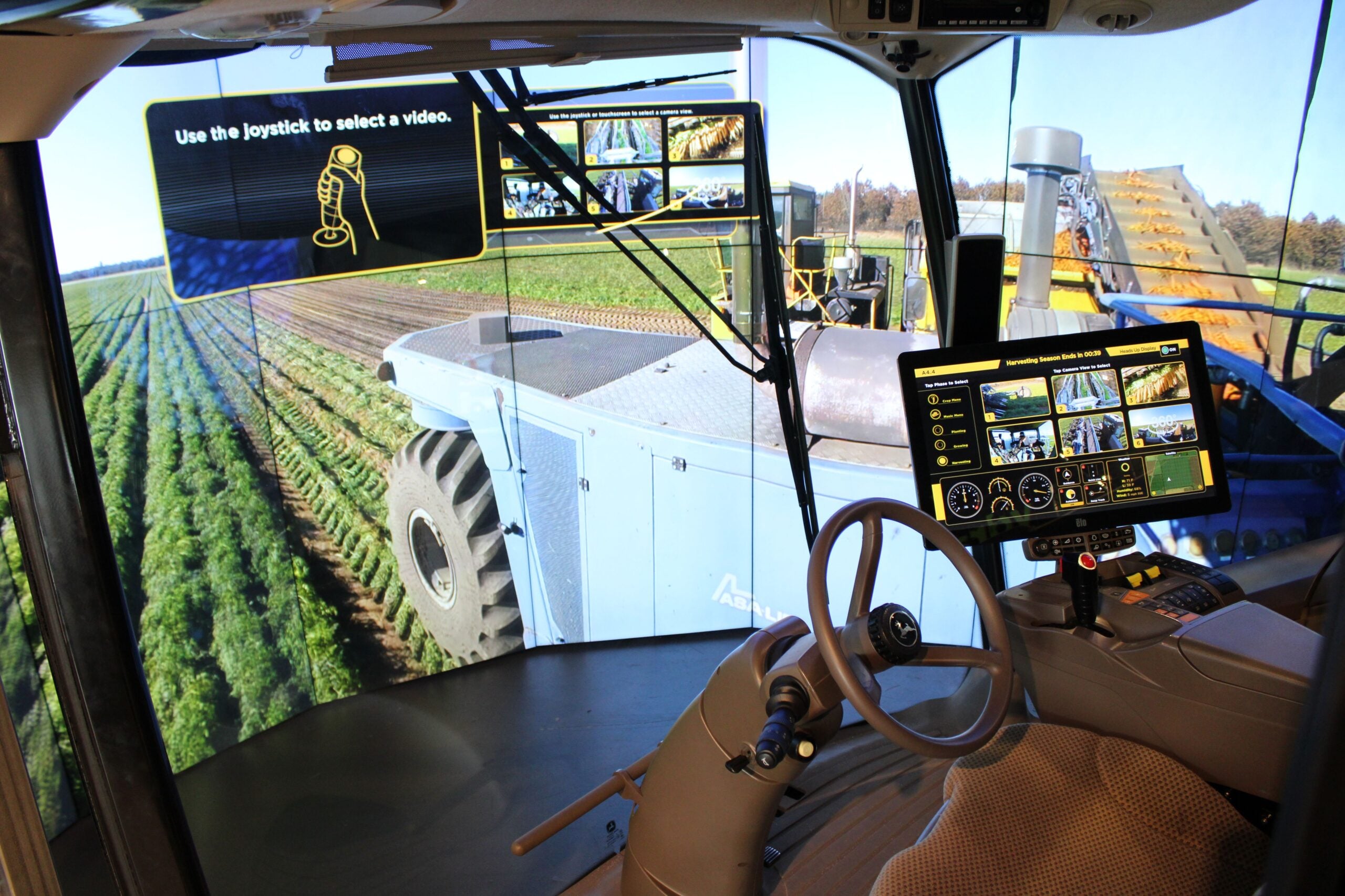View from inside a modern tractor cab with digital controls and screens displaying field footage, joystick instructions, and live camera views of farming equipment operating in a field.