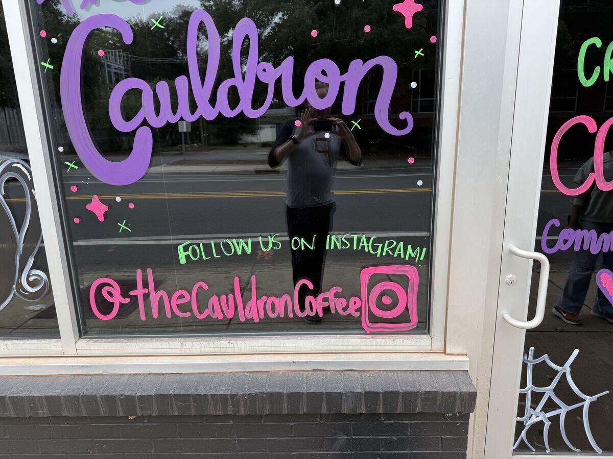 Hand-painted storefront window with large purple lettering that reads “Cauldron” above neon green text that says “FOLLOW US ON INSTAGRAM!” and the handle “@theCauldronCoffee” in bright pink with a painted Instagram logo.