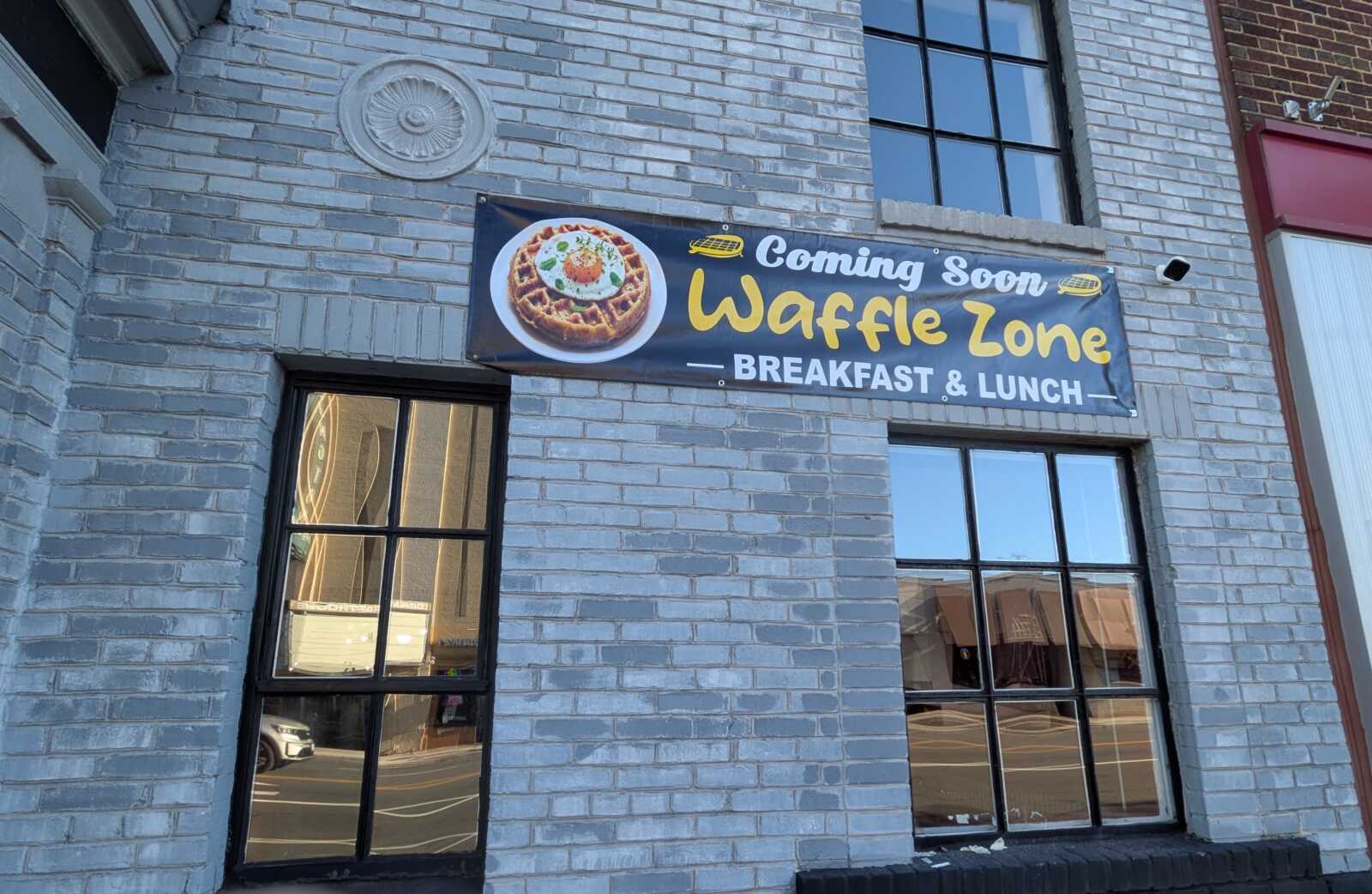 Banner on a gray brick storefront reading 'Coming Soon Waffle Zone' with a waffle image, advertising breakfast and lunch.