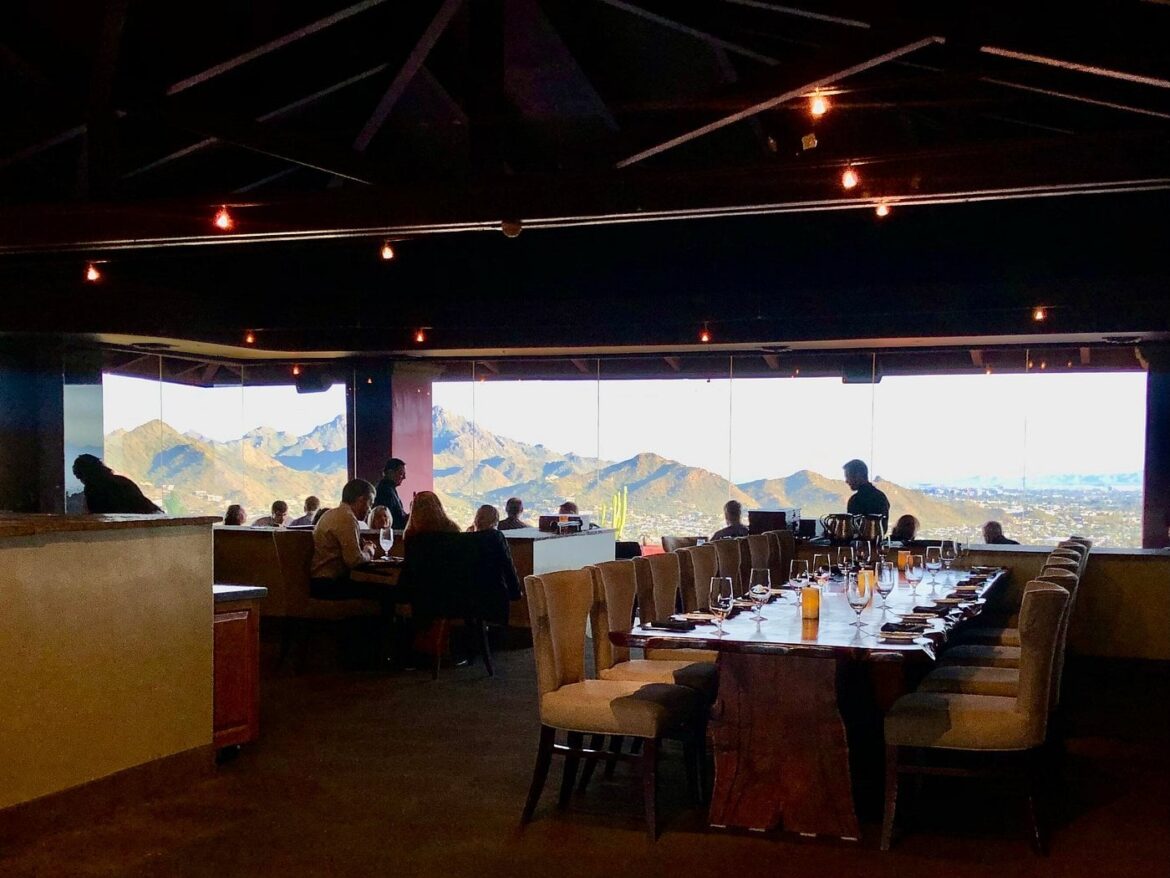Diners seated at tables in a restaurant overlooking a desert mountain landscape at sunset.