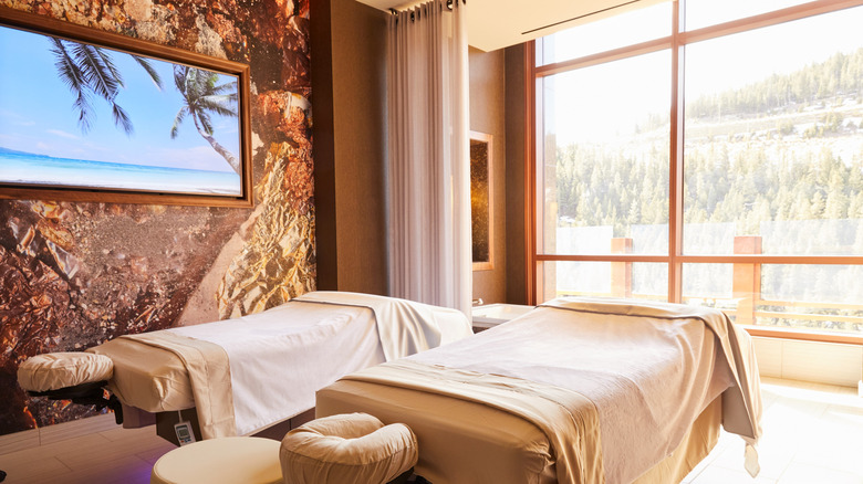 A spa treatment room with two luxurious massage tables facing a wall of windows overlooking mountains covered in evergreen trees