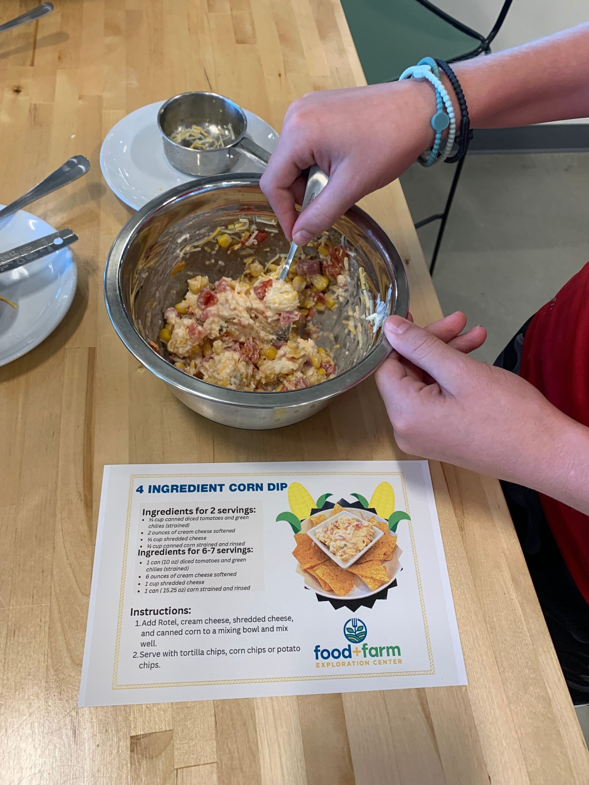 A person mixes corn dip ingredients in a metal bowl next to a printed recipe card titled 4 Ingredient Corn Dip on a kitchen counter.