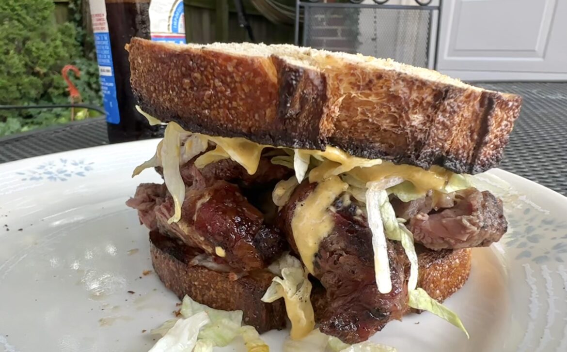 Grilled ribeye steak sandwich on fresh baked sourdough bread with mozzarella cheese, caramelized onions, lettuce, and homemade spicy garlic pesto parmesan mayo.