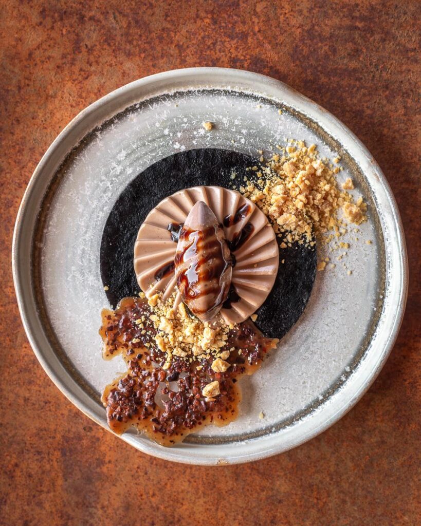 White Chocolate and Cacao Husk Mousse on a Black Cocoa Base, Milk Chocolate Namelaka with Cacao Pulp Reduction, Caramelized White Chocolate Crumble, and Cocoa Nib Tuile
