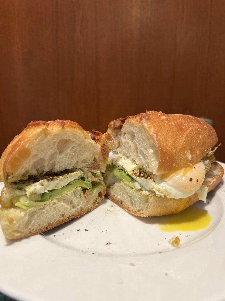 Fried egg cooked with garlic, butter, and Zataar with avocado, pepper, point Reyes truffle toma, and grated parmigiana reggiano on a bacon lardon and Gruyère baguette