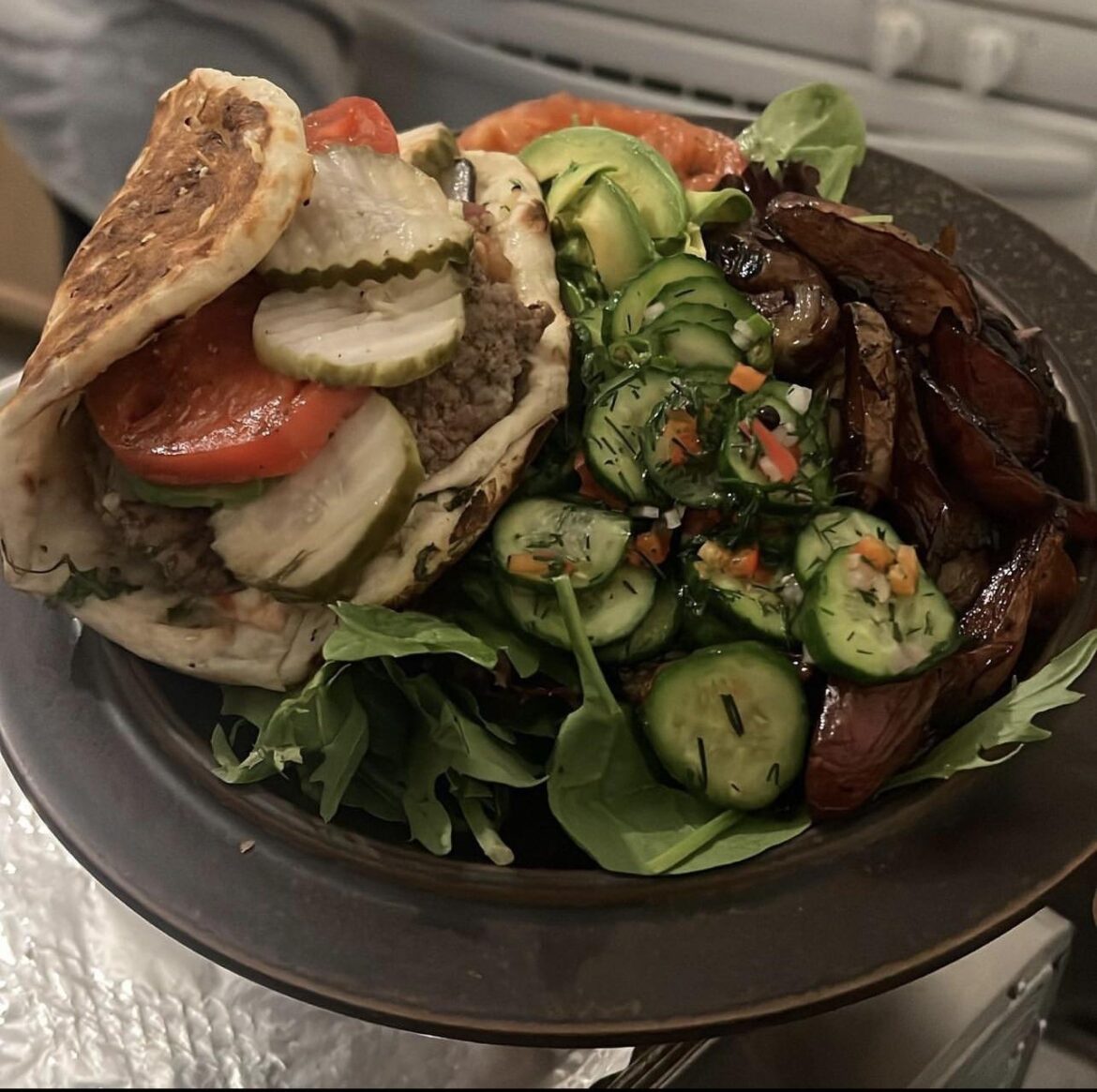 Bacon drunken goat garlic naan burger with a dressed heirloom, pickles, avocado, Hellman mayo, ketchup, and dijon with candied Yuzu dill fermented peppercorn smashed cucumber red pepper Thai chilis garlic shallot salad with fingerling potatoes on a bed of spring mix