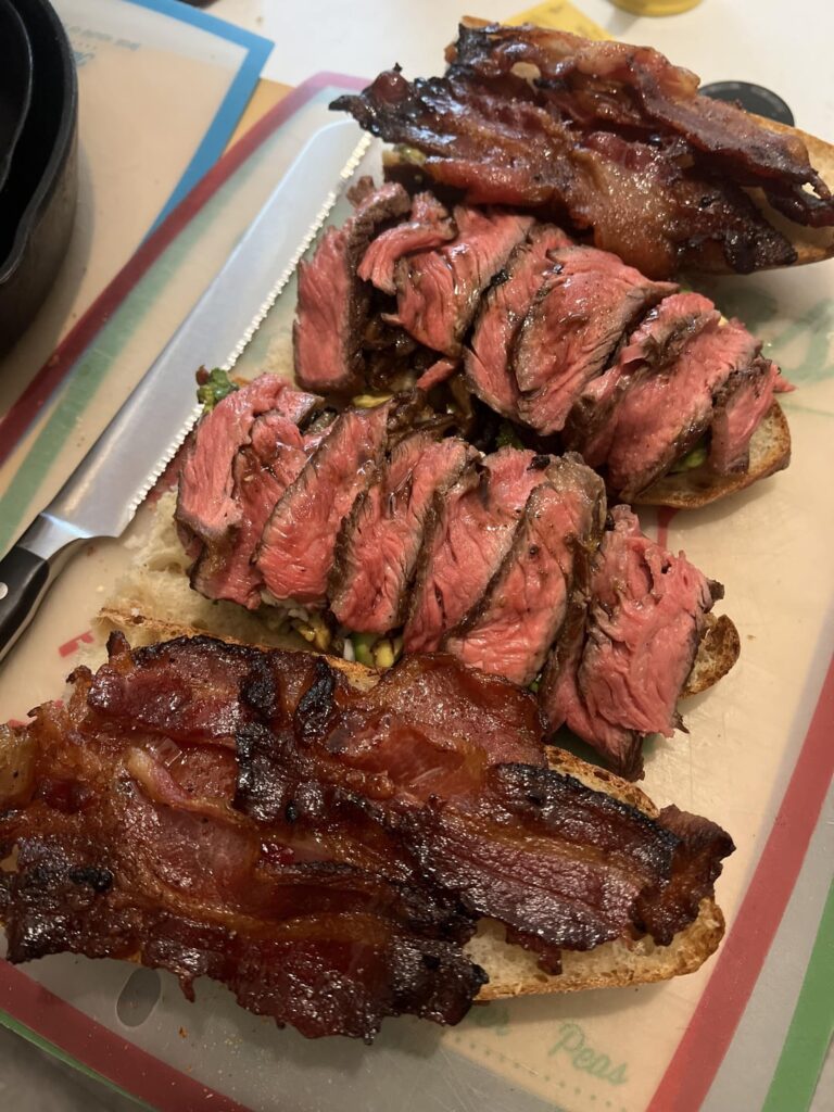 Ribeye and maple bourbon peppered bacon baguette sandwich with baked shiitakes, rogue river blue cheese, sir Kensington mayo, and dressed avocado with date, Thai pepper, shallot, pepper, maldon salt, and cilantro garlic balsamic
