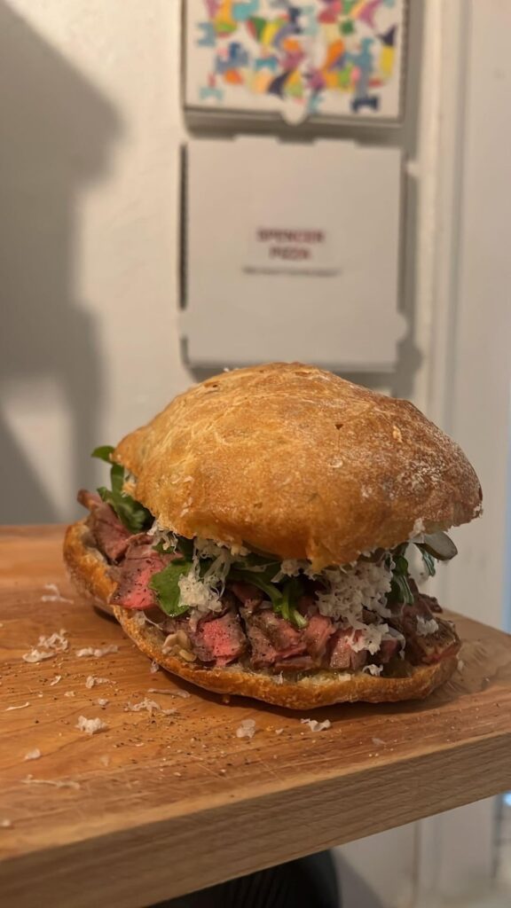 Cold thinly sliced heavily peppered lightly evoo drizzled prime ribeye sandwich on an olive loaf with Jùscht Swiss, honey mustard sauce, dressed arugula and cherry tomatoes