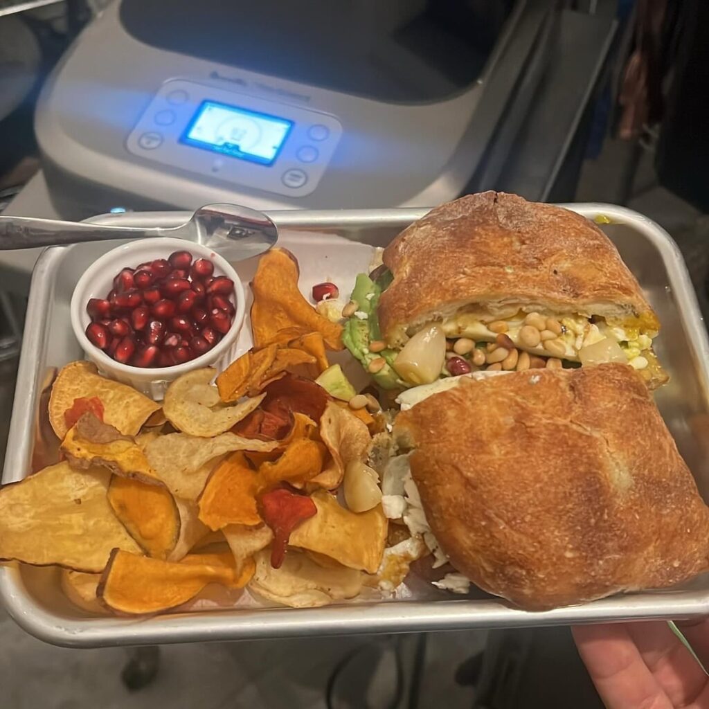 Fried egg sandwich on an olive loaf with avocado dressed in Zataar and pepper, isigny spring butter, smeared roasted garlic, pomegranates, toasted pine nuts, and barrel aged Greek feta with Terra garlic & herb chips and extra pomegranates on the side