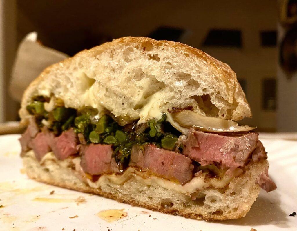 Marinated flank steak with charred broccolini, sautéed oyster mushrooms, caramelized onions, Queso fresco, and truffle mayo on a Portuguese bun.