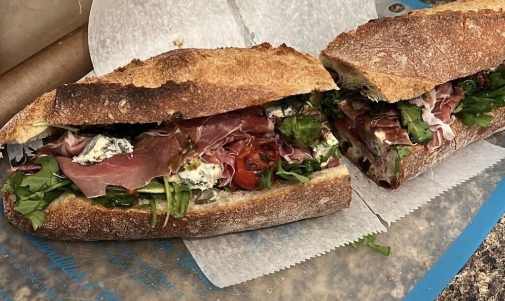 san daniele prosciutto sandwich with dressed arugula, sautéed cherry bomb tomatoes, Giuseppe gran deposito balsamic vinegar, saint agur blue cheese, and sautéed ramp honey butter on an ancienne baguette