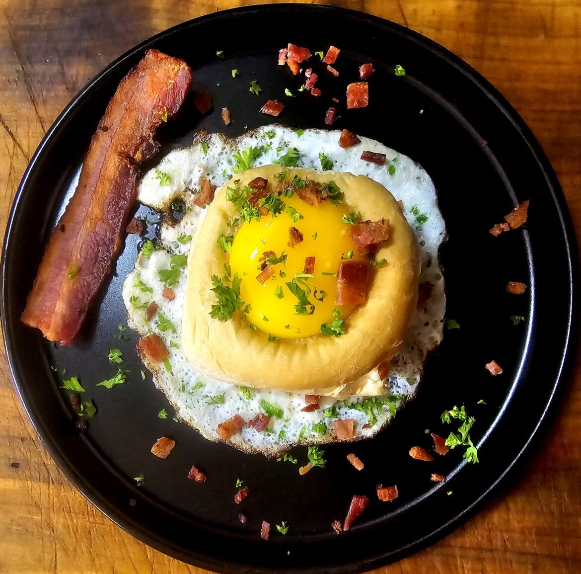 Sausage gravy inside a dinner roll with yolk on top of a fried egg white with scallions and some bacon