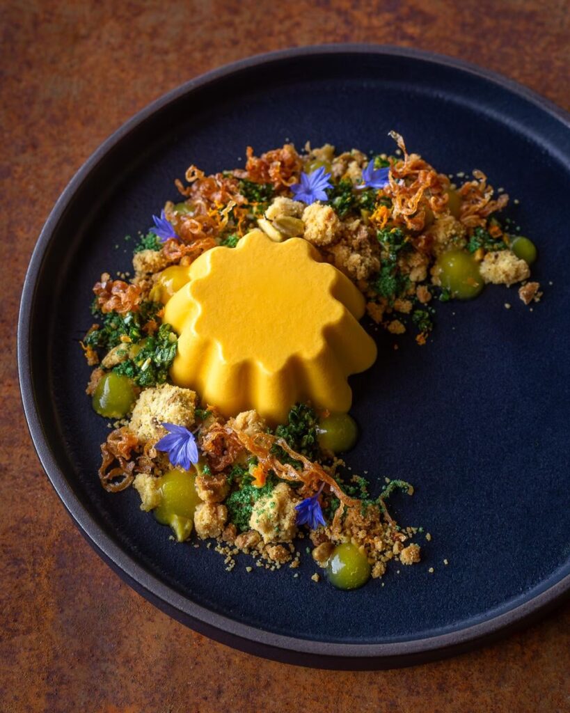 Brown Butter Carrot Panna Cotta With Savory Pistachio Crumble, Brown Butter Powder, Carrot Top Orange Gremolata, Fried Shallots, and Orange Gel