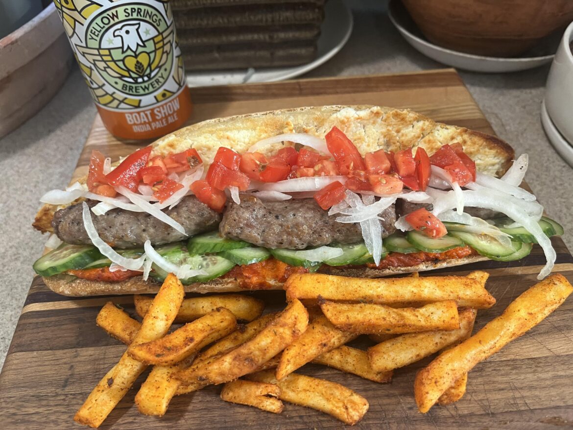 Grilled ćevapčići (Balkan pork, beef, lamb, sausage) with sliced onion, cucumber, and diced tomatoes on a broiled hot ajvar and sheep’s milk cream cheese baguette.