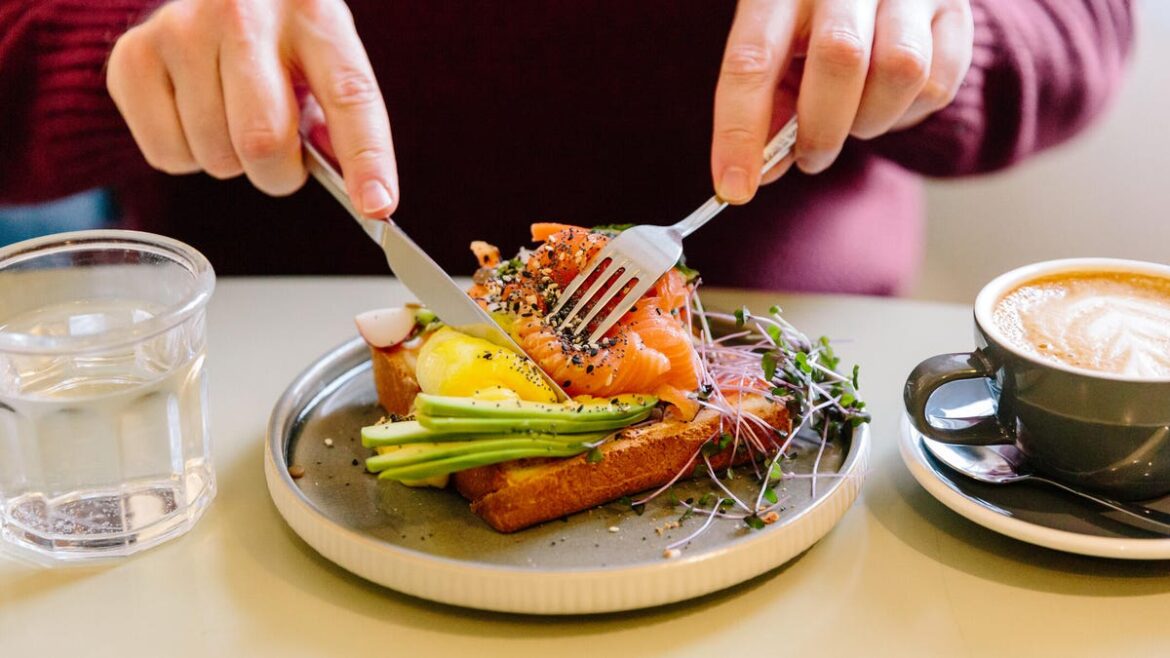 Do You Really Need to Eat 3 Meals a Day? We Asked an Expert Dietician A man's hands cutting a salmon and avocado flatbread with a knife and fork.