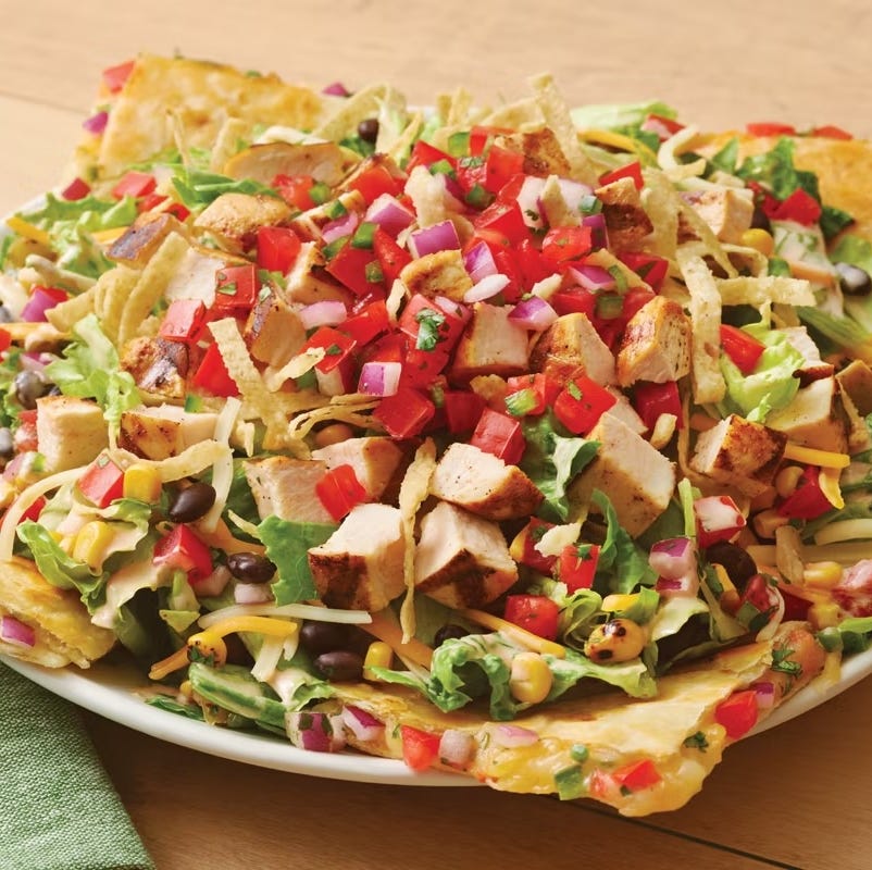 a large salad featuring greens grilled chicken and various toppings served on tortilla chips