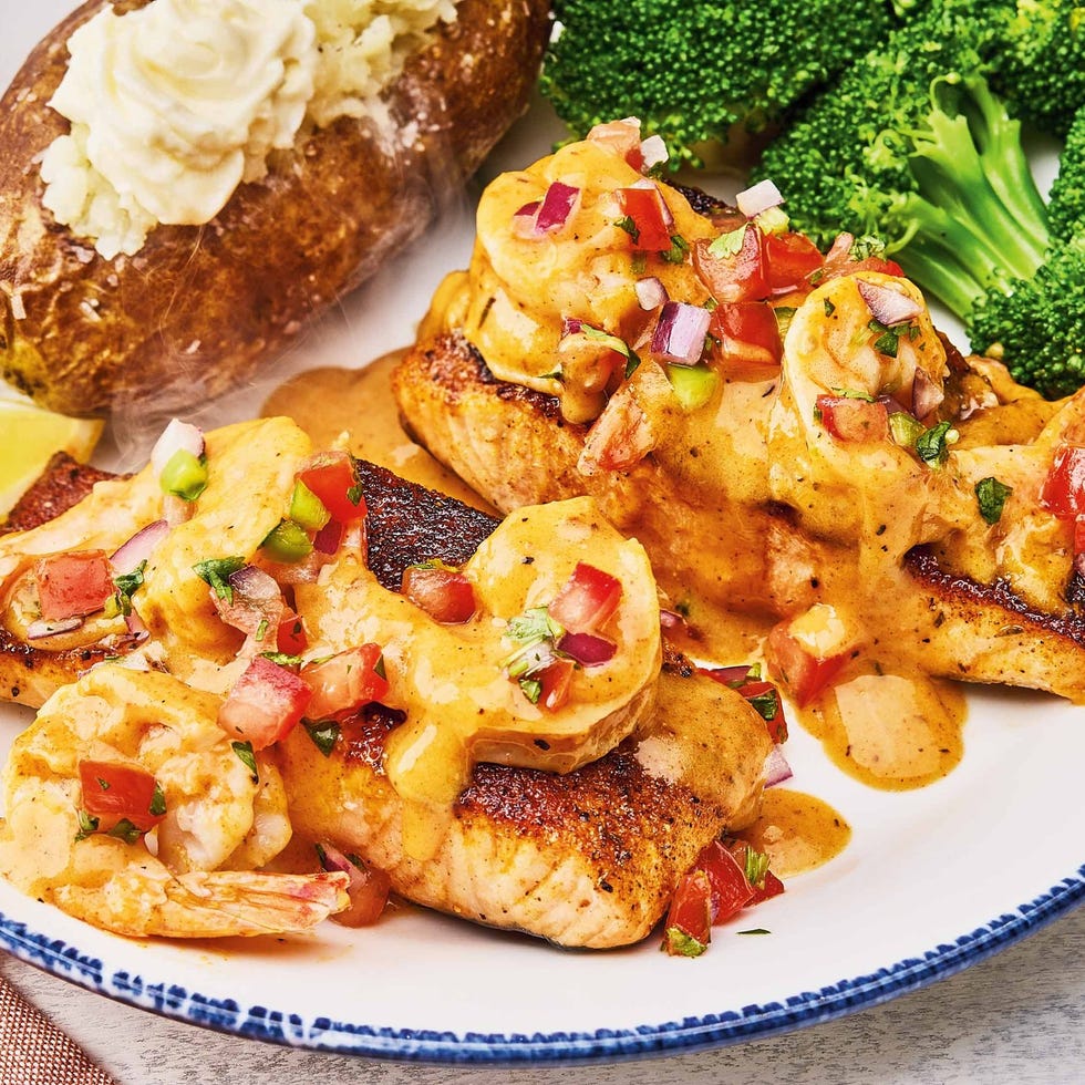 plated dish featuring seasoned fish topped with shrimp and sauce alongside a baked potato and broccoli