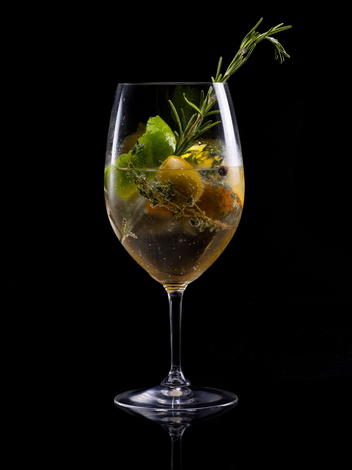 In a wine glass, a gin and tonic is served with olives and a sprig of rosemary.