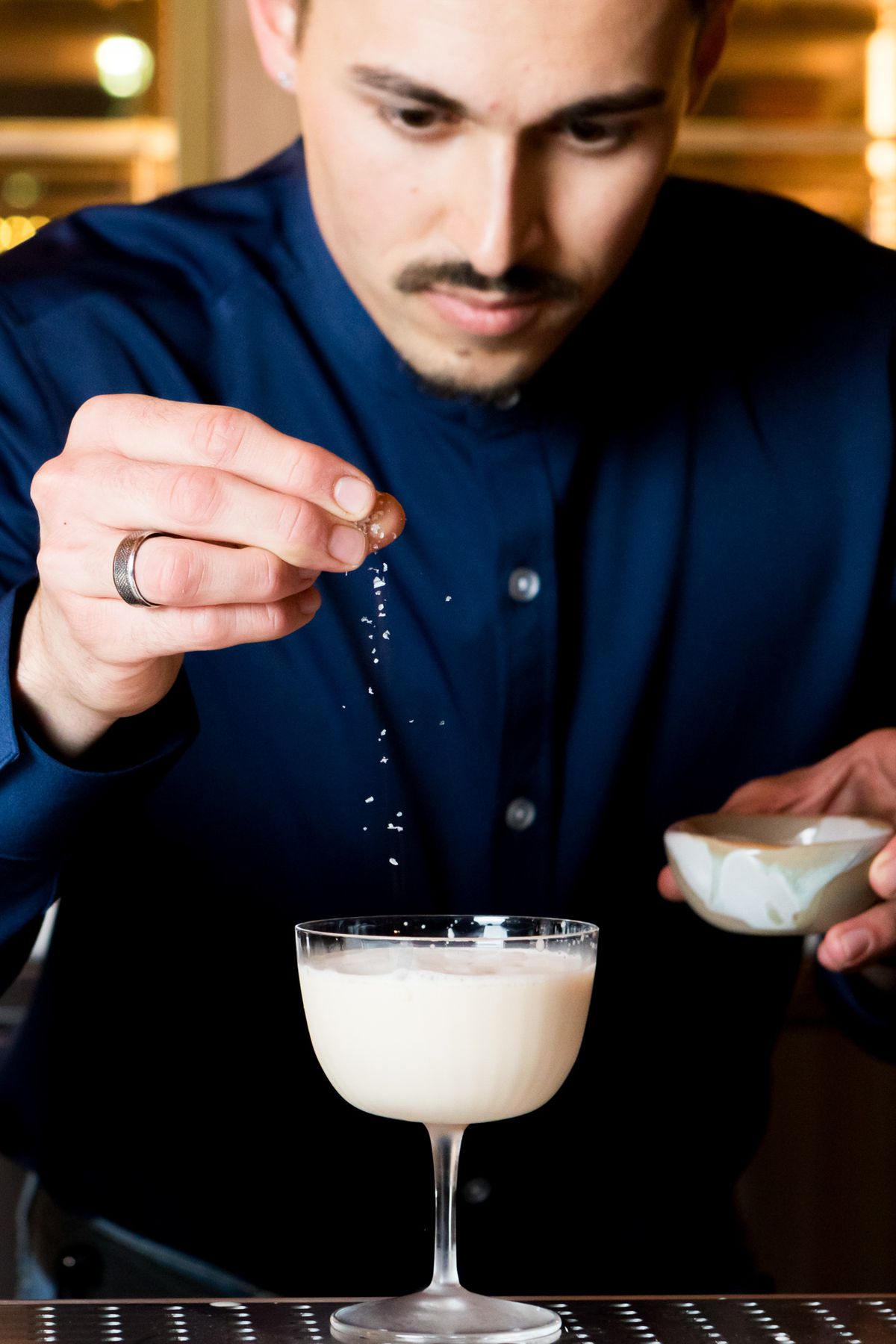 A man sprinkles salt over a white, foamy cocktail in a coup glass. 