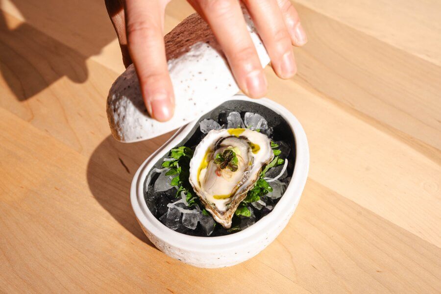 A hand lifts the lid of a white ceramic dish containing a garnished oyster on ice, an offering from one of Houston's hottest new restaurants, all elegantly set on a light wooden surface.