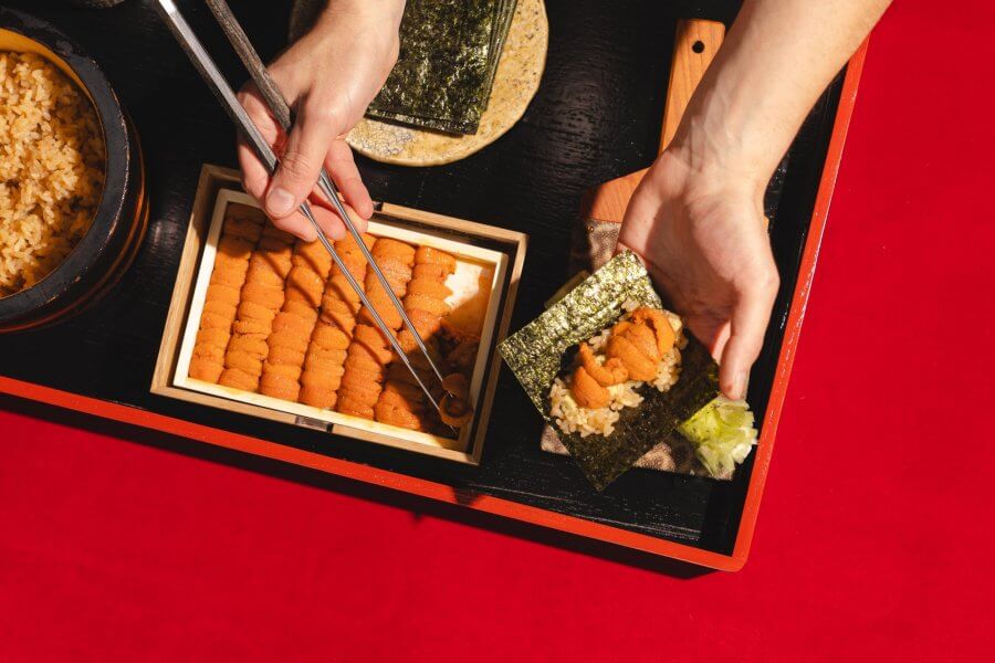 Hands delicately prepare uni sushi on a tray with nori, rice, and wooden boxes of sea urchin roe. 